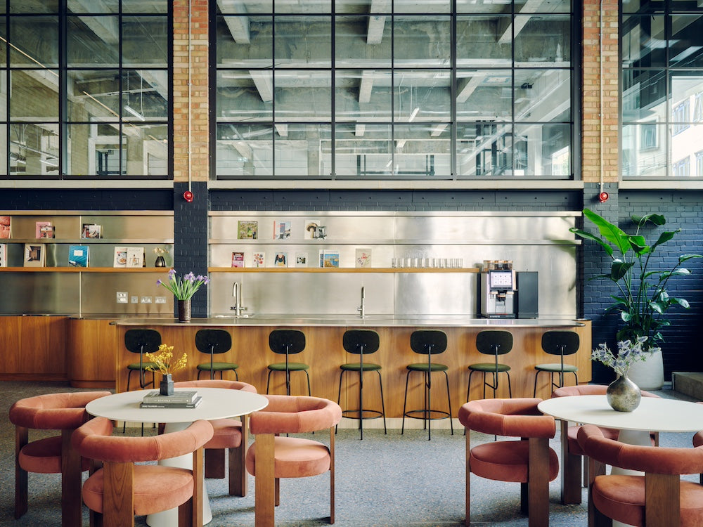 A modern industrial-style cafe or lounge interior with round tables, blush pink upholstered chairs, a wood counter, black bar stools, large black-framed windows, exposed ceiling pipes, brick columns, and potted plants.